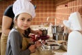 Lovely little girl in chef's hat and kitchen apron, smiling, looking at camera, helping her mother to prepare yummy Royalty Free Stock Photo