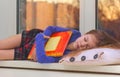 Caucasian little girl of 5 years sleeping hugging big book on windowsill with sunset view in window