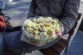 Caucasian male in a winter jacket, holding out a southwestern salad in a plastic to-go container at an outdoor table