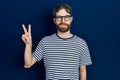 Caucasian man with beard wearing striped t shirt and glasses smiling with happy face winking at the camera doing victory sign Royalty Free Stock Photo