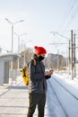 caucasian man in a dark jacket and red hat at a train station waiting for a train and texting on his smartphone Royalty Free Stock Photo