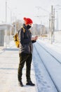 caucasian man in a dark jacket and red hat at a train station waiting for a train and texting on his smartphone Royalty Free Stock Photo