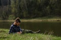 Caucasian man with dreadlocks and beard lying in clearing on river bank in spring park and working on laptop. Serious and