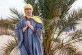 Caucasian man dressed in dishdasha and yellow headscarf standing in desert against palm tree, an European tourist