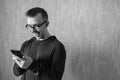 Caucasian man in glasses looks at phone and rejoices on gray background. Monochrome portrait of delighted male with copy space.