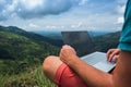 Caucasian man with laptop sitting on the edge of ella mountain with stunning views of the valley in Sri Lanka.