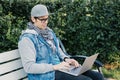 A Caucasian man sits on a park bench and works remotely through a laptop