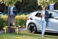Caucasian man standing near an electric car that is charging and making time adjustments on a smartphone. Plug charging