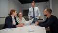 A caucasian man stands and holds a drawing, three colleagues sit at a table and listen to him.