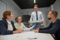 A caucasian man stands and holds a drawing, three colleagues sit at a table and listen to him.