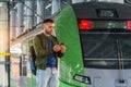 Caucasian man traveler in green windbreaker and with backpack stands on rail road platform typing text on phone, electric commuter