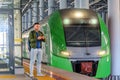 Caucasian man traveler in green windbreaker and with backpack stands on rail road platform typing text on phone, electric commuter Royalty Free Stock Photo