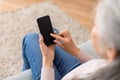 Caucasian mature gray-haired female typing on smartphone with empty screen in living room