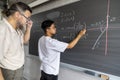 Caucasian mature man teacher supervises teen asian high school boy student writing maths exercise on blackboard with chalk