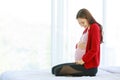 Caucasian millennial young happy female prenatal pregnant mother model in casual pregnancy outfit jacket sitting alone on bed in Royalty Free Stock Photo