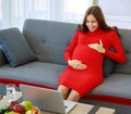 Caucasian millennial young happy sexy female prenatal pregnant mother in casual red pregnancy dress sitting on sofa holding hands Royalty Free Stock Photo