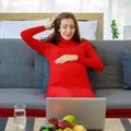 Caucasian millennial young happy sexy female prenatal pregnant mother in casual red pregnancy dress sitting on sofa holding hands Royalty Free Stock Photo