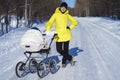 Caucasian mom in yellow coat is walking with white baby carriage on snow road among the forest at winter sunny day Royalty Free Stock Photo