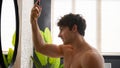 Caucasian naked man guy male in bathroom morning beauty routine procedure looking at mirror reflection apply spray hair