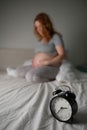 Caucasian pregnant woman sits on the bed and suffers from insomnia. Alarm clock in the foreground. Royalty Free Stock Photo