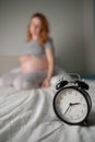 Caucasian pregnant woman sits on the bed and suffers from insomnia. Alarm clock in the foreground. Royalty Free Stock Photo