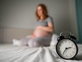 Caucasian pregnant woman sits on the bed and suffers from insomnia. Alarm clock in the foreground. Royalty Free Stock Photo