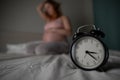 Caucasian pregnant woman sits on the bed and suffers from insomnia. Alarm clock in the foreground. Royalty Free Stock Photo