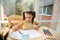 Top view. Preschooler girl in a yellow T-shirt and two ponytails, enjoying watercolor painting during art class at home