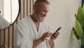 Caucasian relaxed senior old man in bathroom in bathrobe calm mature retired male in bathroom browsing mobile phone