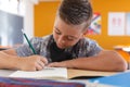 Caucasian schoolboy with face mask sitting in classroom concentrating and writing in schoolbook