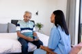Caucasian senior man holding medicines talking with biracial female physiotherapist in bedroom