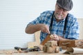 Caucasian senior old white bearded man carpenter in apron and hat using chisel working in workshop, tools machine small wooden toy Royalty Free Stock Photo