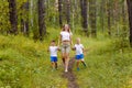 A Caucasian slender smiling woman and two cheerful preschoolers children in white t-shirts run along the path, holding hands in