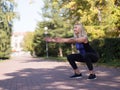 Caucasian smiling middle-aged athletic woman doing deep squat on footway in urban park, outdoor, selective focus Royalty Free Stock Photo