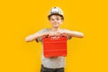 Caucasian teenage boy wears protective helmet and glasses hold instrument case on yellow background. Child like builder or worker