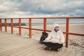 Caucasian woman in hat and jacket with backpack in winter sits on wooden pier on beach near North Sea. Denmark Copenhagen tourist Royalty Free Stock Photo