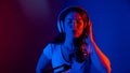 Caucasian woman in headphones singing into a microphone in neon light on a black background. An emotional girl is Royalty Free Stock Photo