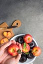 Caucasian woman holds in hands vivid ripe red apple with stem green leaves dark plums on white enamel plate. Wooden cutting board Royalty Free Stock Photo