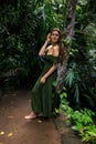 Caucasian woman leaning against a tree in tropical rain forest. Beautiful woman wearing long green dress. Walking trail in jungle Royalty Free Stock Photo