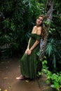 Caucasian woman leaning against a tree in tropical rain forest. Beautiful woman wearing long green dress. Walking trail in jungle Royalty Free Stock Photo