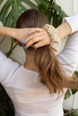 Caucasian woman with long natural blond hair making ponytail using beige scrunchie. Warm, positive tones. Against monstera plant Royalty Free Stock Photo