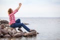 Caucasian woman sitting on stones on sea and making selfie with smartphone, copy space with water Royalty Free Stock Photo