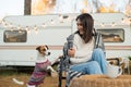 Caucasian woman sitting in a wicker chair wrapped in a blanket with a dog in the yard near the trailer in autumn.