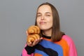 Caucasian woman wearing sweater posing isolated over gray background, holding delicious croissant, smells with pleasure, likes Royalty Free Stock Photo