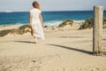 Caucasian woman with white dress viewed from back walking alone on the sand to the beach and blue ocean - water and sky in horizon Royalty Free Stock Photo