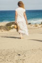 Caucasian woman with white dress viewed from back walking alone on the sand to the beach and blue ocean - water and sky in horizon Royalty Free Stock Photo