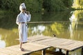 Caucasian Woman In White Robe And Towel Is Fishing Holding Spinning Rod On Pier Of Lake