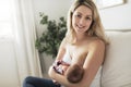 Woman in her bedroom on the white cushions, smiling and breastfeed her baby.