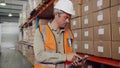 Caucasian worker typing on digital tablet assorting parcels according to checklist standing in factory warehouse