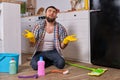 Caucasian young bearded man sits on the floor of his kitchen and tries to clean it using all of his detergents, rags